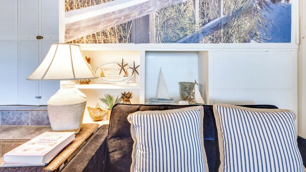 A cozy living room with a sofa, a lamp, a book, and a decorative shelf with a painting and small items. - Valdosta Home: Bar, Bbq, Fire Pit + More! (Valdosta)