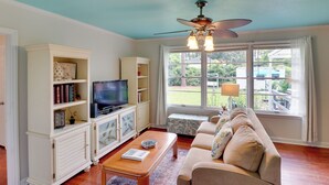 A living room with a television, bookshelves, a sofa, and a window with a view of the outdoors. - Valdosta Home: Bar, Bbq, Fire Pit + More! (Valdosta)