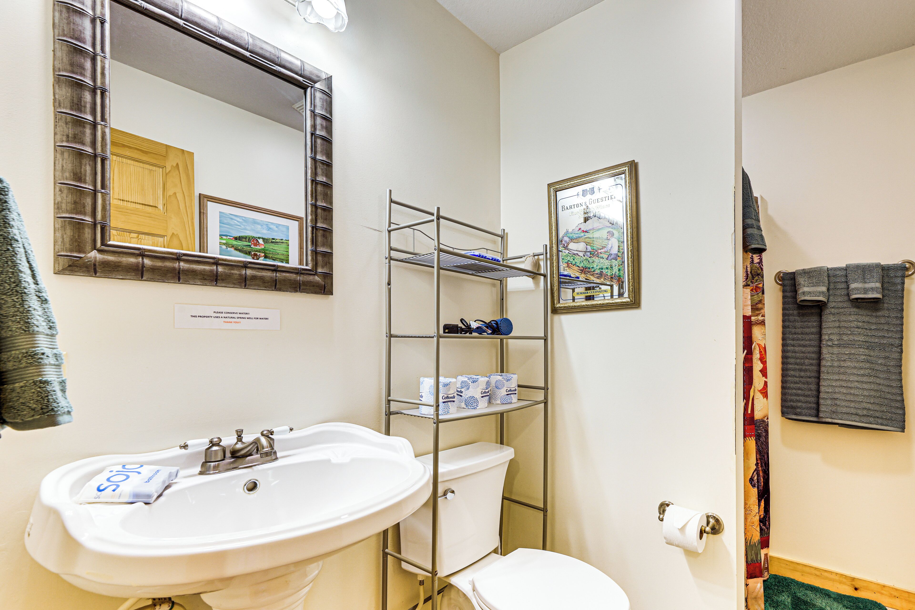 A bathroom with a white sink, a toilet, a metal shelf with toiletries, a framed picture, and a towel rack.