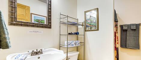 A bathroom with a white sink, a toilet, a metal shelf with toiletries, a framed picture, and a towel rack.