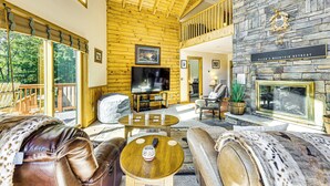 A spacious living area with a stone fireplace, wooden ceiling, and large windows offering a view of the outdoors. - Deck & Mtn Views: Log Cabin By Snowshoe Mountain! (Slaty Fork)