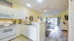 A modern kitchen with white cabinetry, a built-in oven, and a dishwasher. There is a dining area with a table and chairs, and a view of the outdoors through a large window. - 2 Mi to Beach: Grand Strand Condo w/ Pool Access! (North Myrtle Beach)