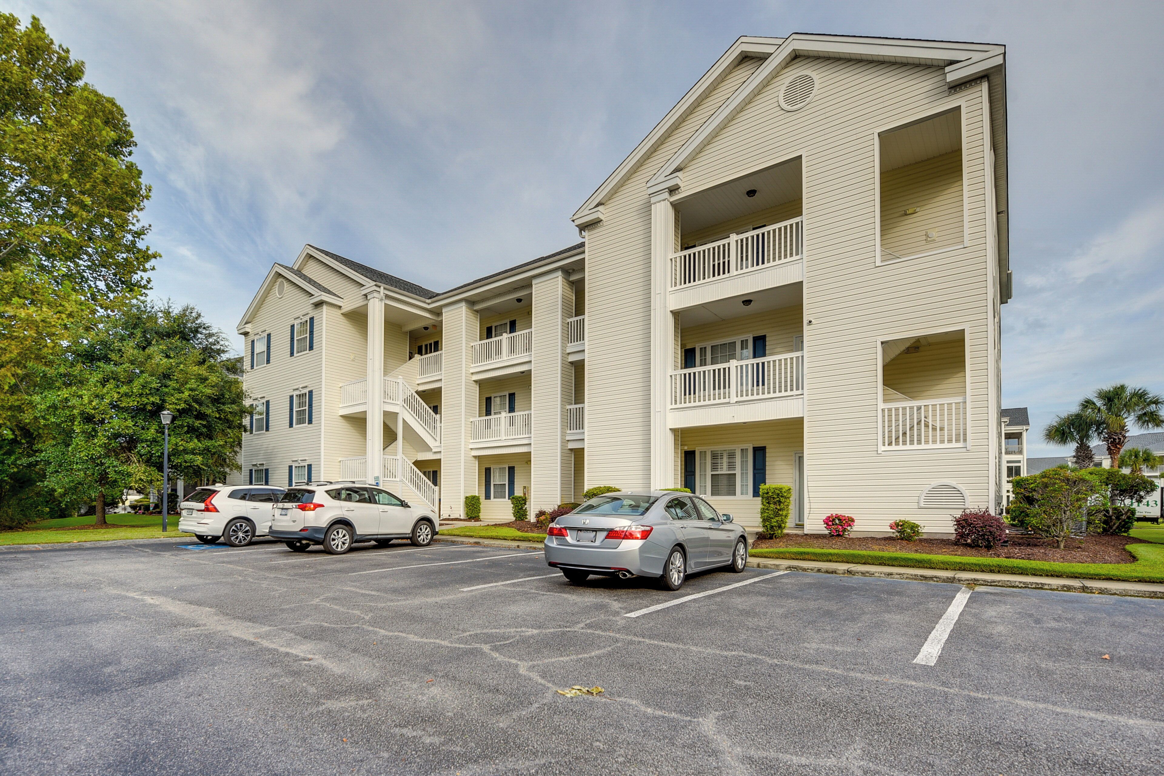 A multi-story residential building with balconies, a parking lot, and a clear sky.
