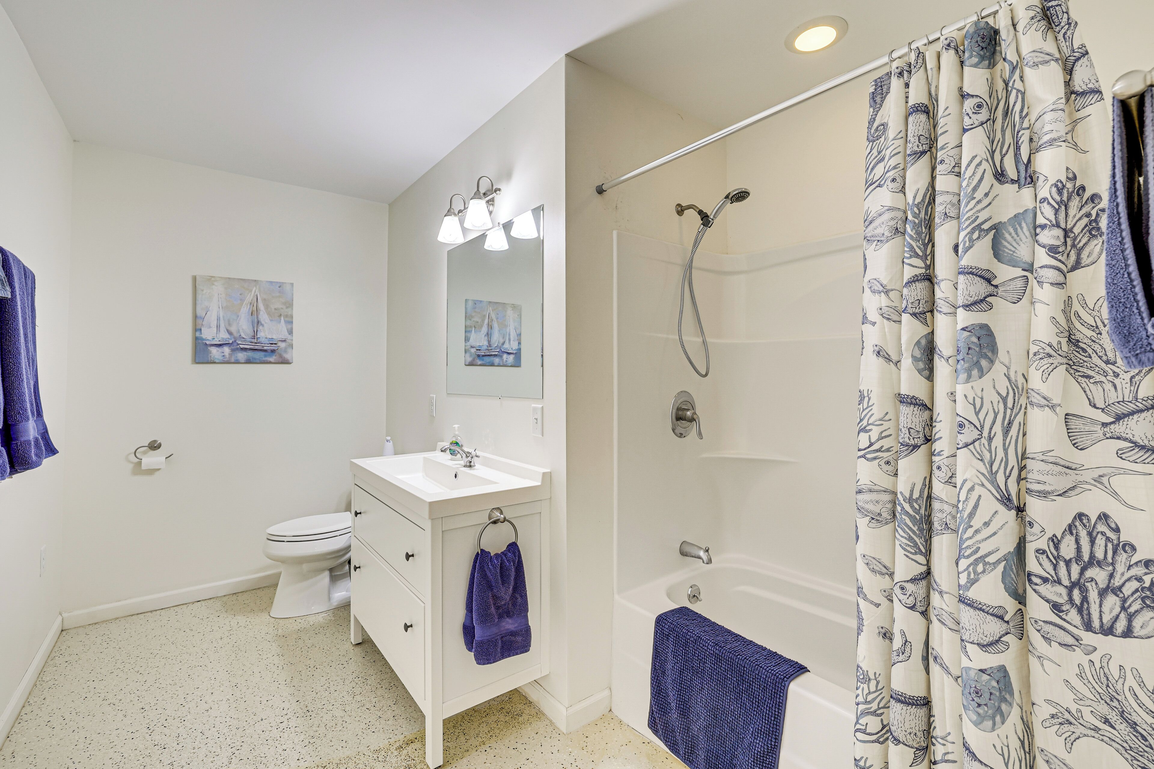 A bathroom with a white sink, toilet, and bathtub. There are two paintings on the wall, a towel rack with a blue towel, and a shower curtain with a marine design.