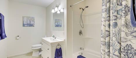 A bathroom with a white sink, toilet, and bathtub. There are two paintings on the wall, a towel rack with a blue towel, and a shower curtain with a marine design.