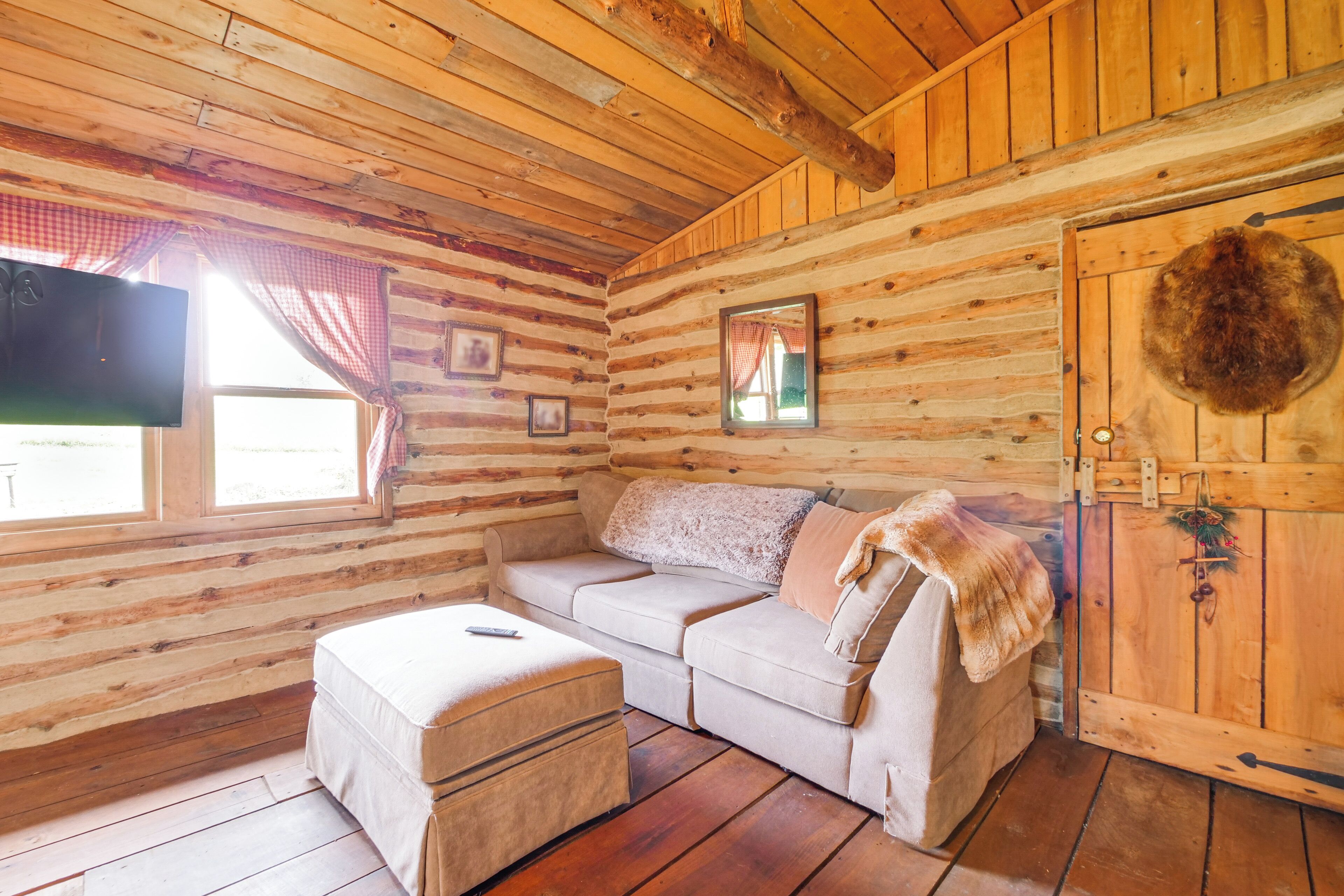 A log cabin interior with a sofa, ottoman, wooden walls, a mounted television, and a door with a fur decoration.