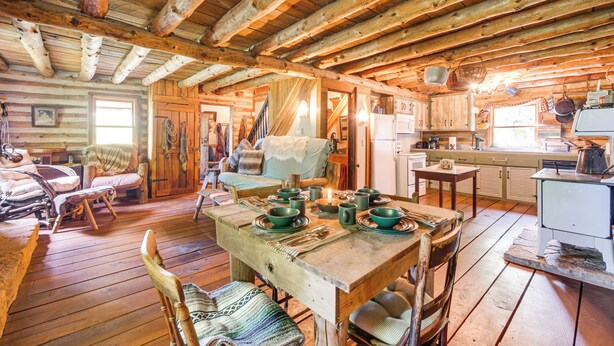 A rustic cabin interior with a wooden dining table set for four, a cozy seating area with a sofa and armchairs, and a kitchen area with basic appliances. - 1800s Romantic Cabin! Fireplace + 3 Jacuzzi Tubs (Paris)