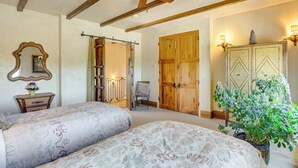 Une chambre avec deux lits, des poutres apparentes en bois au plafond, un ventilateur de plafond, une grande armoire en bois, une plante en pot et un miroir posé sur une commode.