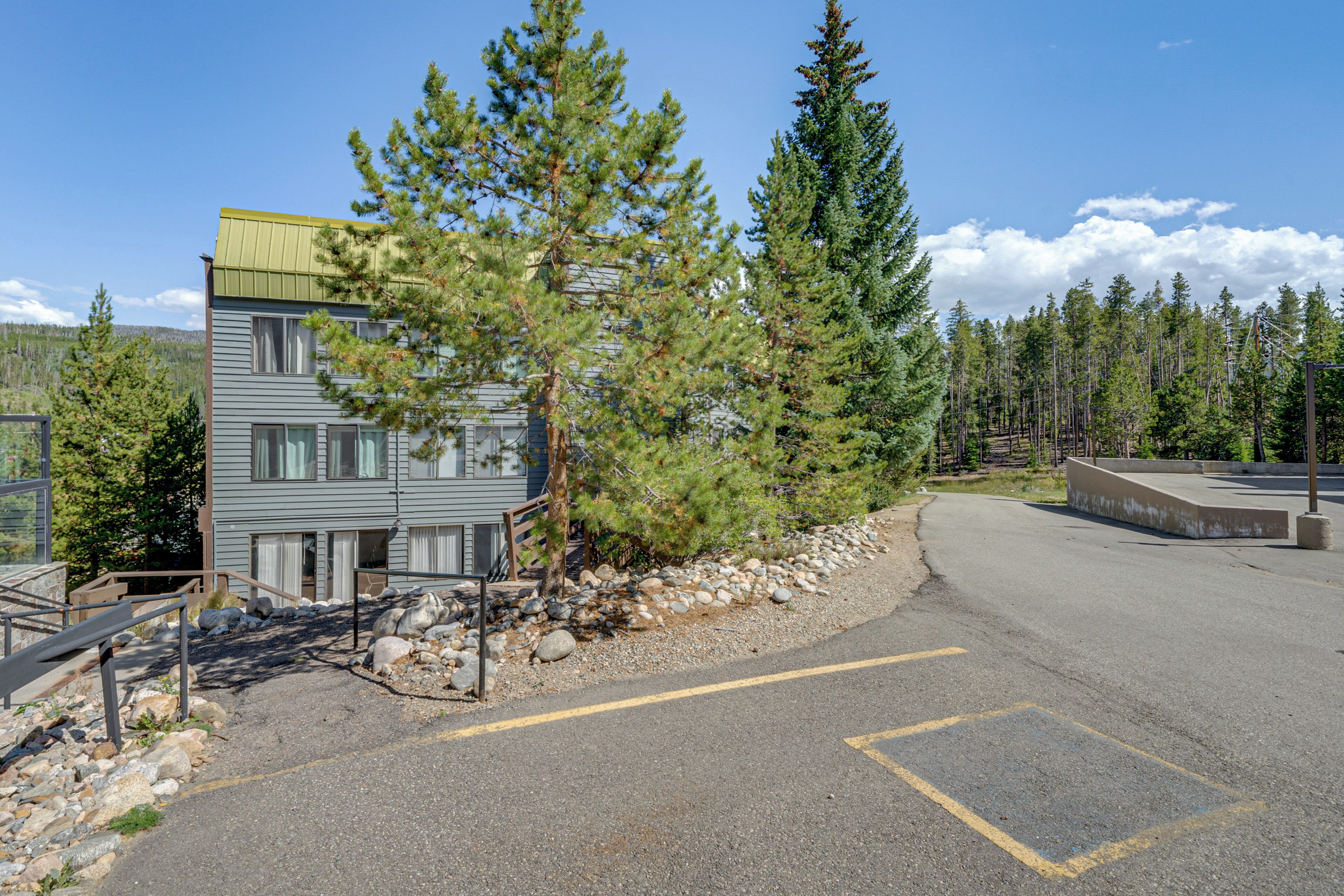 A building with a grey exterior and a green roof, surrounded by trees and a gravel path.