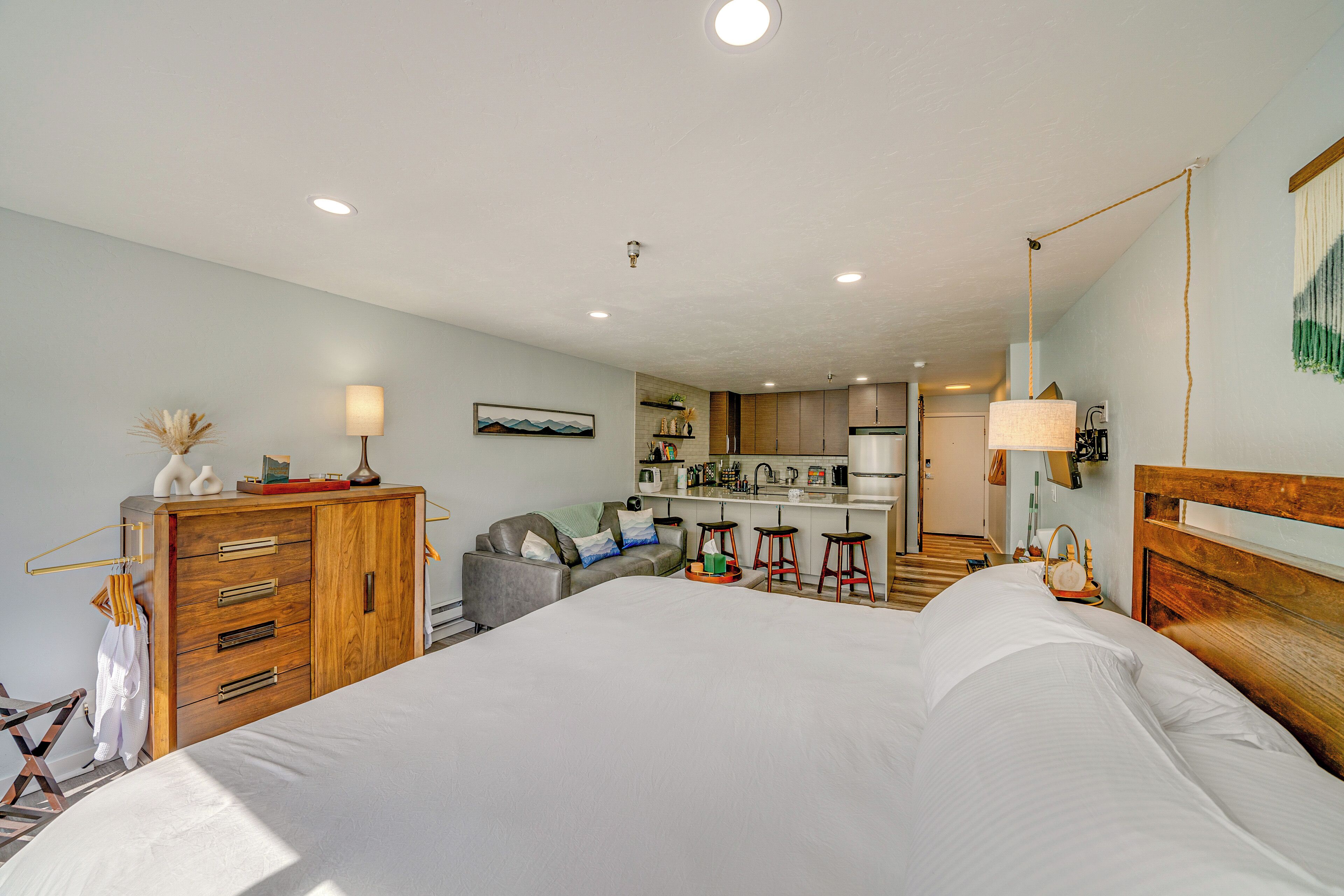 A modern hotel room with a large bed, a wooden dresser, a desk, a kitchenette, and a bathroom.