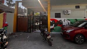Une entrée fermée par un portail, avec une moto garée devant, une voiture rouge garée à droite et un bâtiment sur lequel est peinte une fresque en arrière-plan.