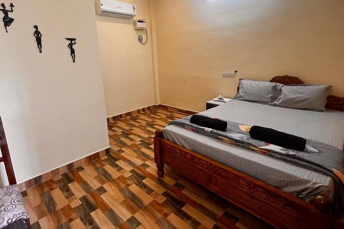 A bedroom with a wooden floor, a bed with grey bedding, a ceiling fan, and an air conditioning unit on the wall.