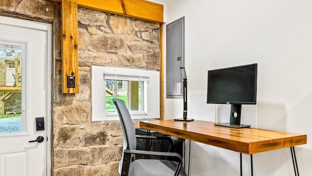Ein Zimmer mit Steinwand, einem Holztisch, einem schwarzen Bürostuhl, einem Computermonitor und einer Tür mit Fenster.