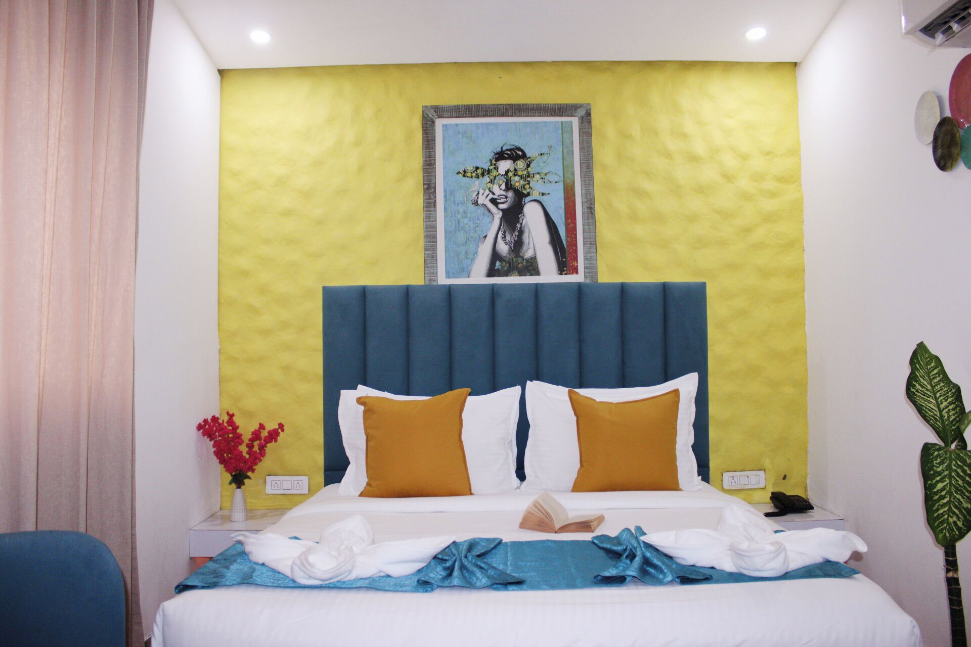 A hotel room with a bed, a painting on the wall, a plant, and a vase with flowers.
