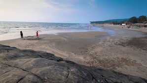 A beach with rocky foreground, calm sea, and distant shoreline with buildings and trees. - Royale Amata By the Beach (Morjim)