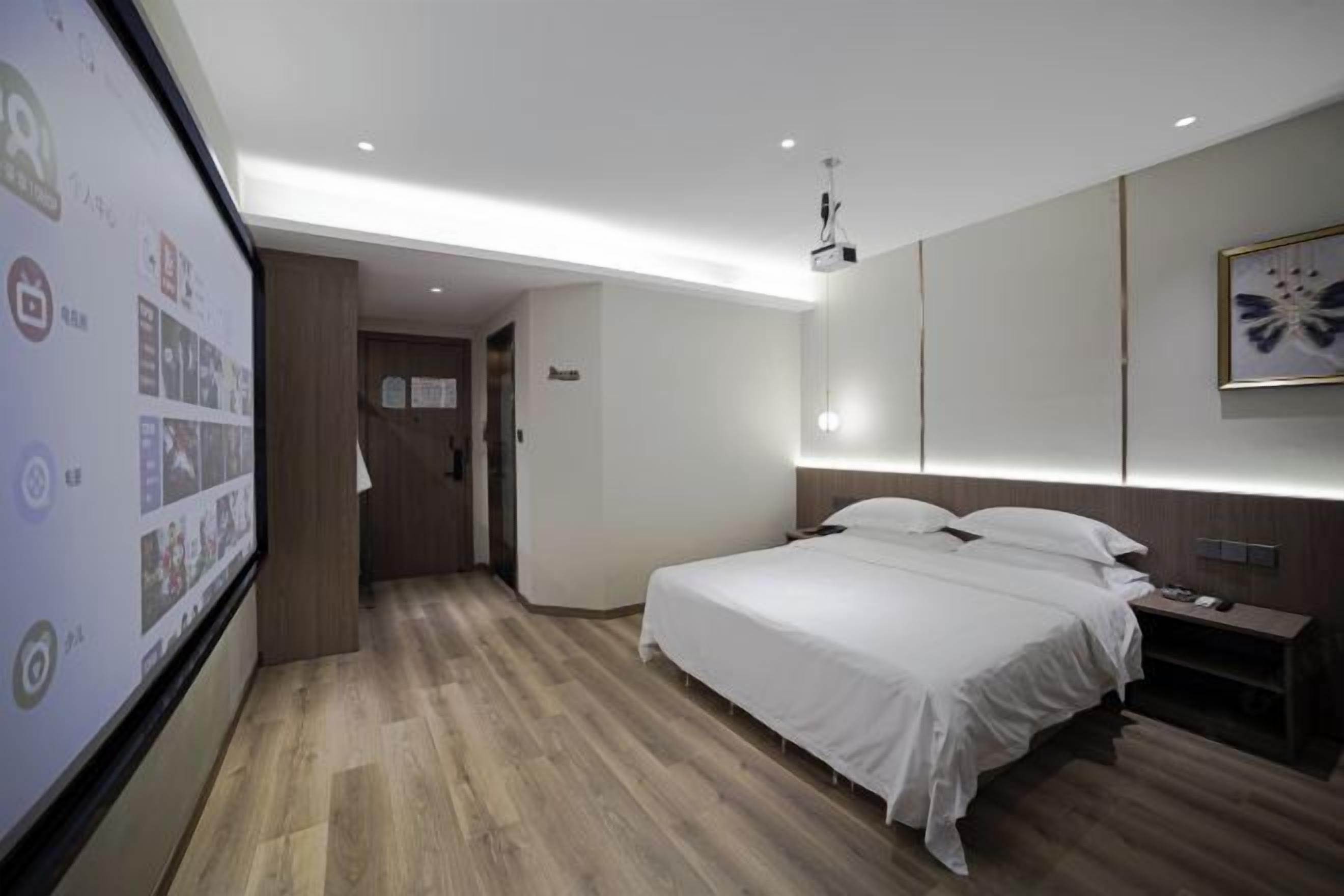 Quarto de hotel moderno com uma cama grande, uma mesinha de cabeceira, uma luminária e uma TV fixada na parede, além de um projetor de teto.