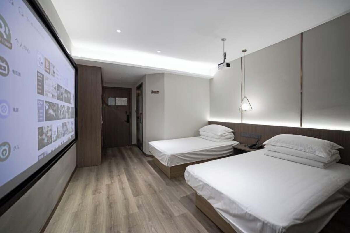 A hotel room with two beds, a large screen displaying images, and a wooden floor.