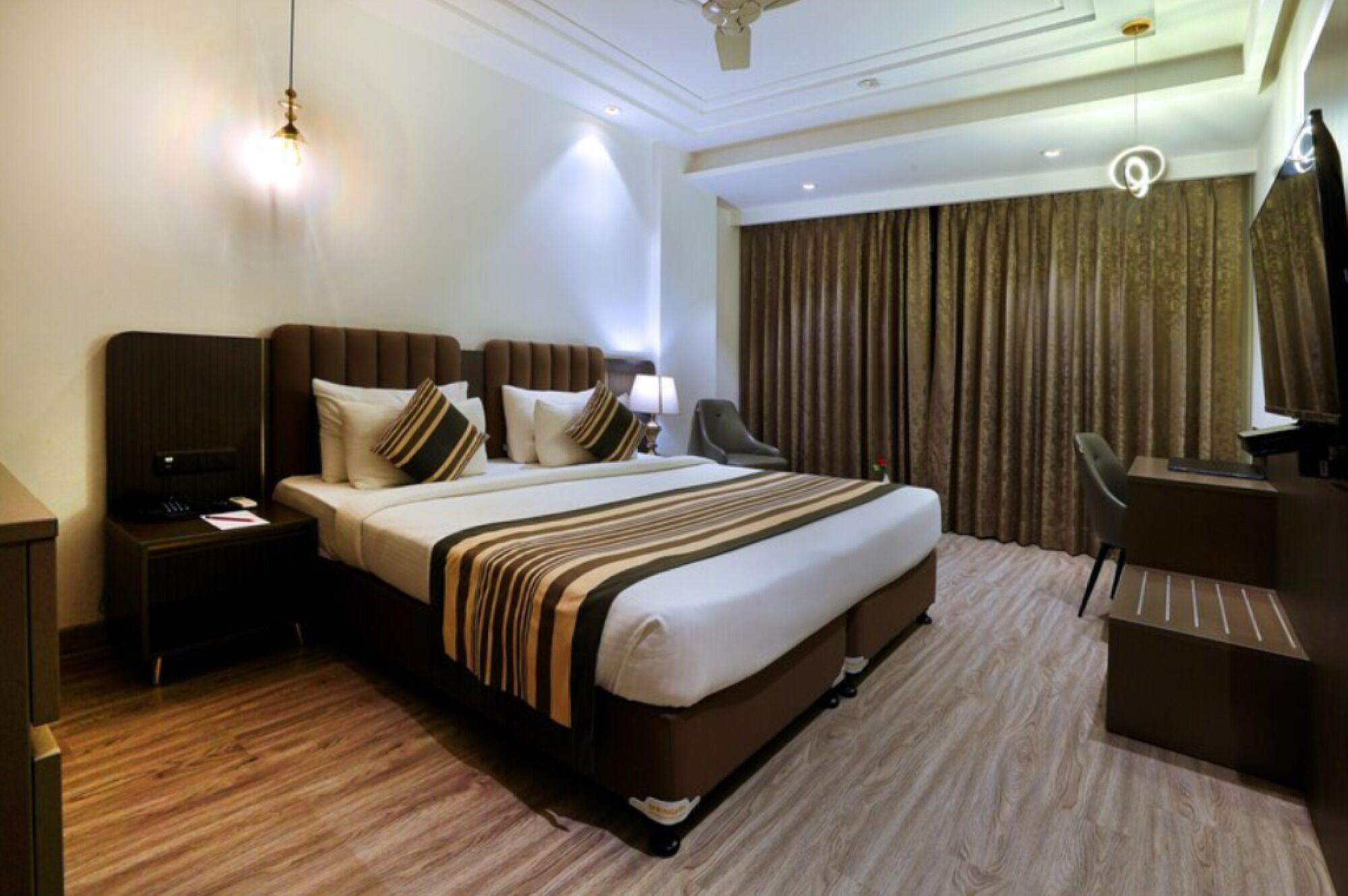 A hotel room with a large bed, a desk, a chair, a TV, and a ceiling fan.