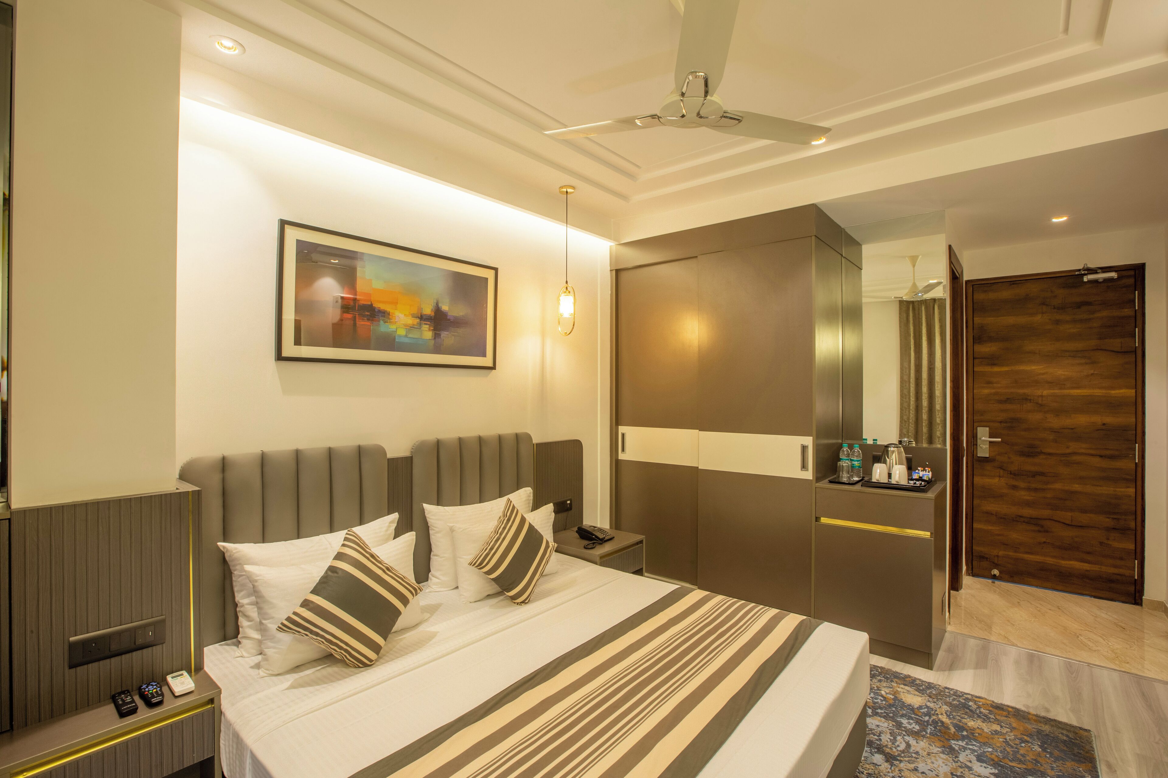A modern hotel room with a large bed, bedside tables, a nightstand, a desk, a wardrobe, a ceiling fan, and a painting on the wall.
