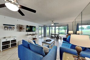 A spacious living area with a ceiling fan, a flat-screen TV, a dining table with chairs, and a sofa with a lamp.