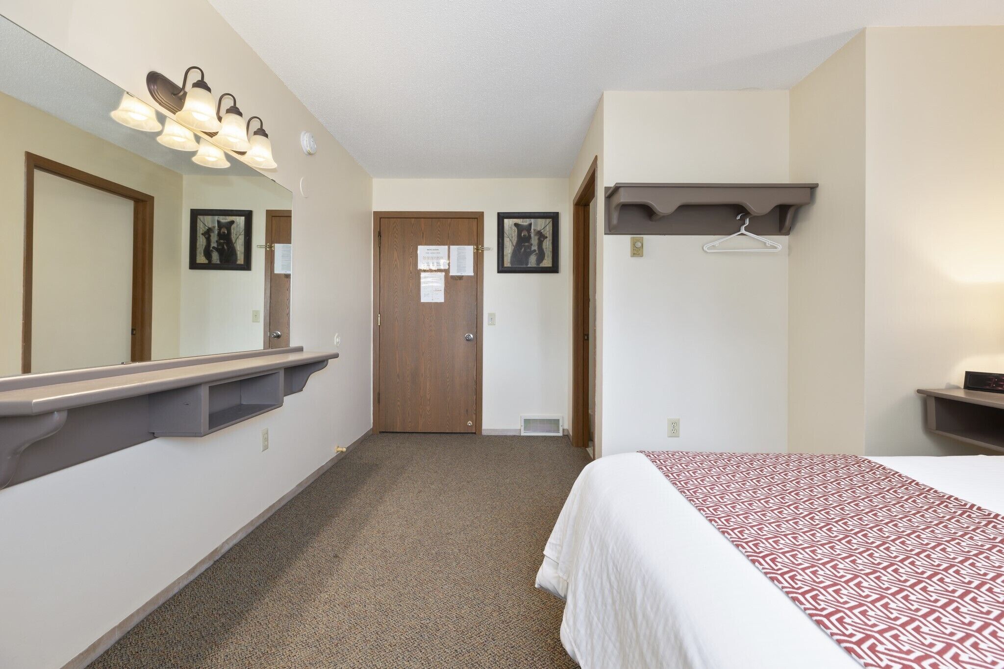 Una habitación de hotel con una cama, un espejo, una lámpara, una puerta y un perchero.