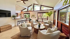 A spacious living area with high ceilings, a ceiling fan, a kitchen area with a bar, and large windows offering views of greenery. - Short Walk to Beach Hanalei Bay (Hanalei)