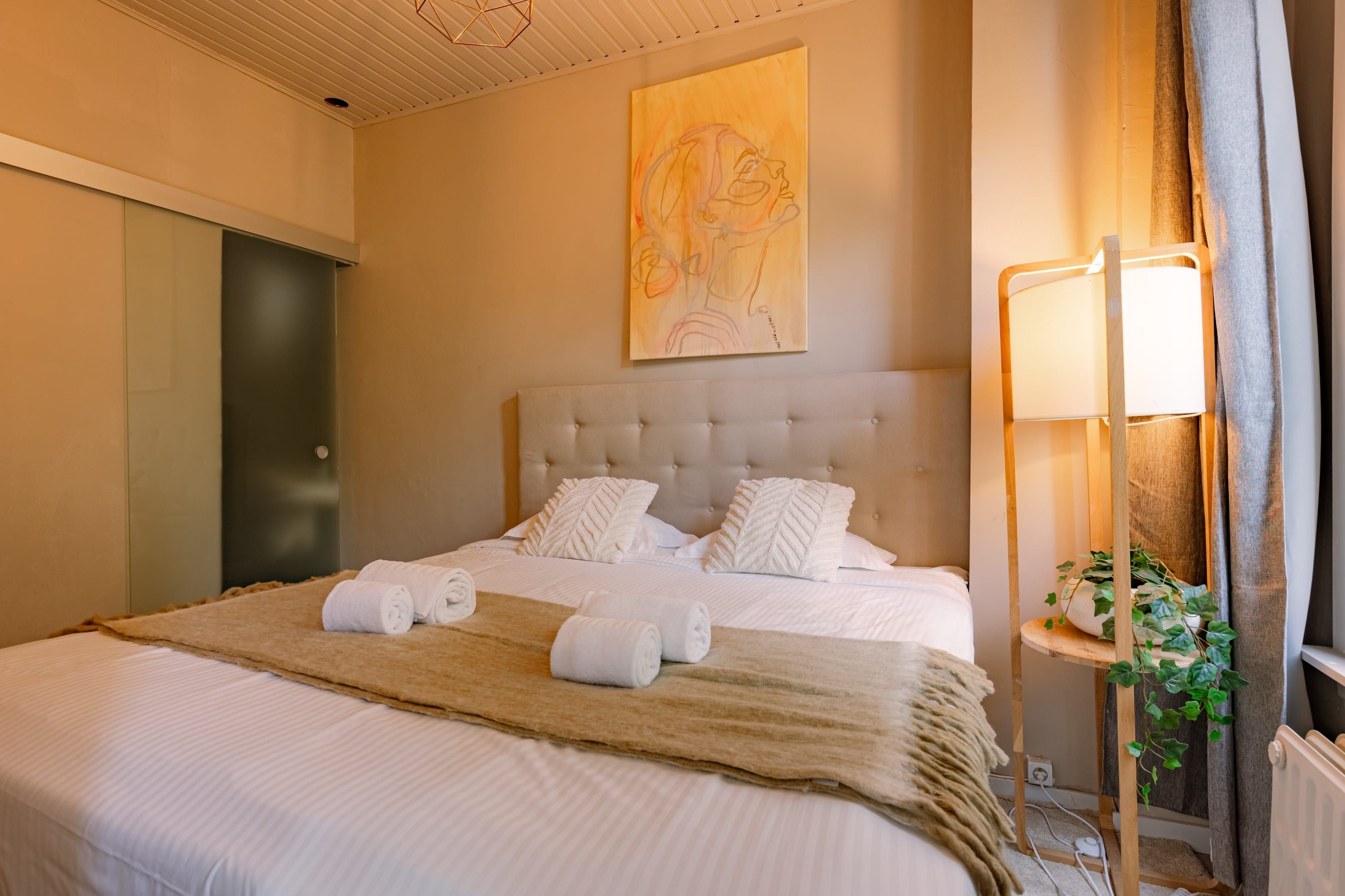 A neatly made bed with white linens and rolled towels, a tufted headboard, a bedside lamp, and a wall art piece.