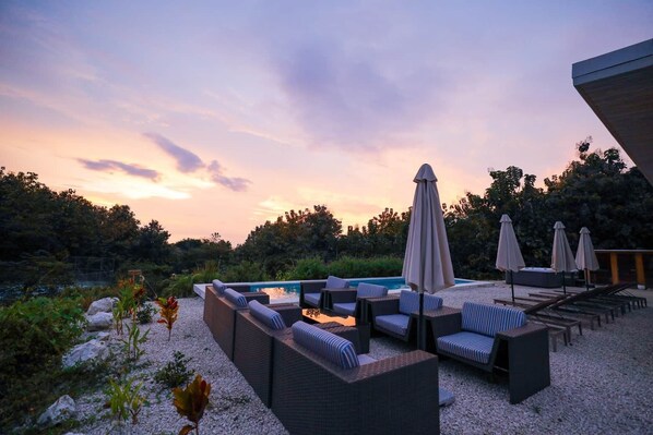 Un área junto a la piscina con sillones de descanso y sombrillas, rodeada de vegetación natural y un cielo despejado.