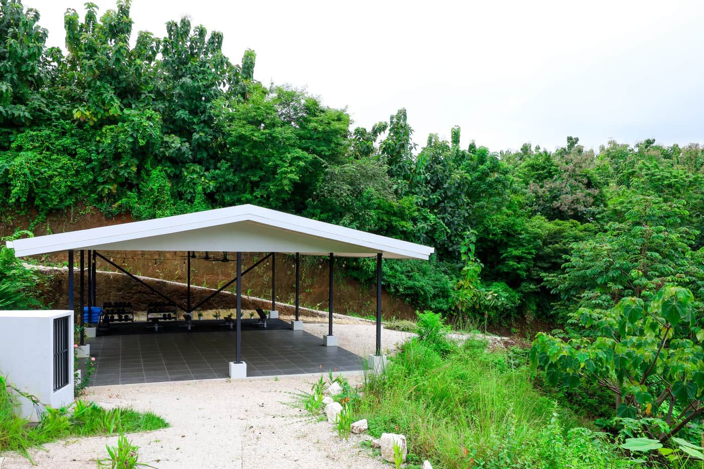 A covered outdoor seating area with a white roof, surrounded by lush greenery.
