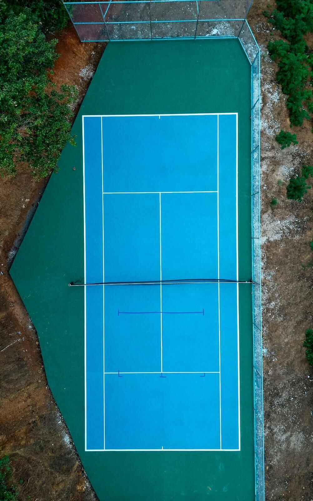 An aerial view of a tennis court with a net in the center, surrounded by green grass and trees.