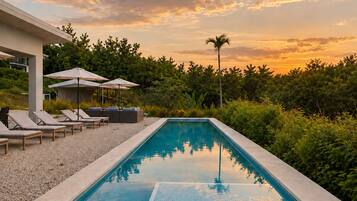 A rectangular swimming pool with a tiled edge, surrounded by lounge chairs and umbrellas, set against a backdrop of lush greenery and a sunset sky.
