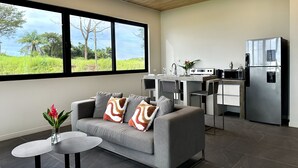Un salon moderne avec un canapé gris, une petite table basse et une vue sur une cuisine équipée d’un réfrigérateur en acier inoxydable et d’un coin repas.