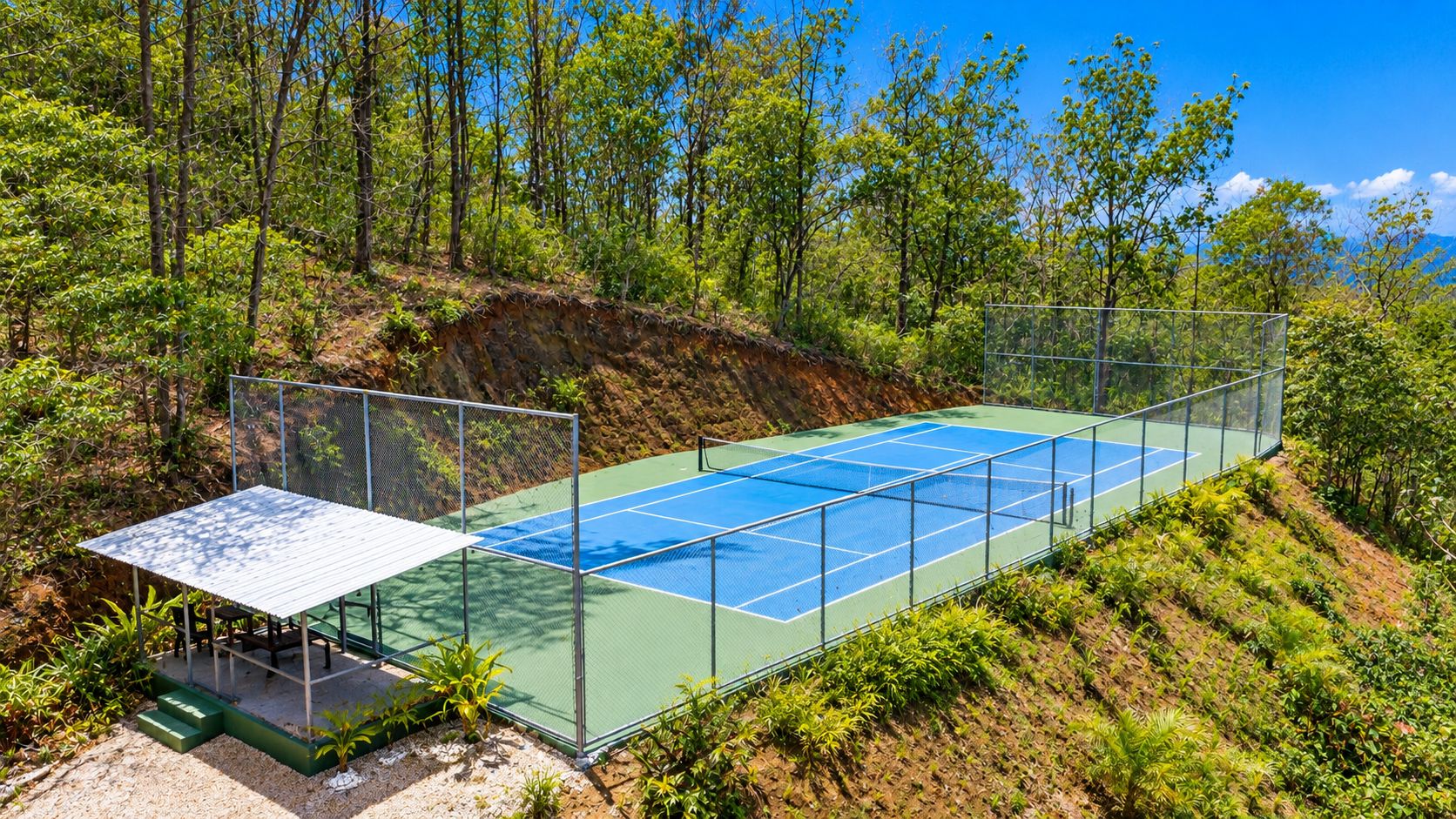 An outdoor tennis court surrounded by a metal fence, with a covered seating area and greenery in the background.
