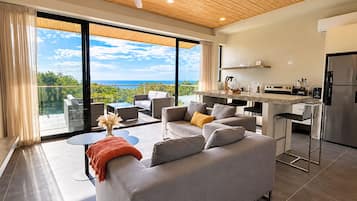 A modern living room with a grey sofa, a coffee table, and a view of the ocean through large windows.