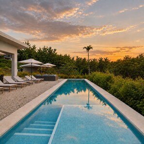 A rectangular swimming pool with tiled edges, surrounded by lounge chairs and umbrellas, set against a backdrop of trees and a sunset sky.