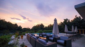 Un área junto a la piscina con sillones de descanso y sombrillas, rodeada de vegetación natural y un cielo despejado.
