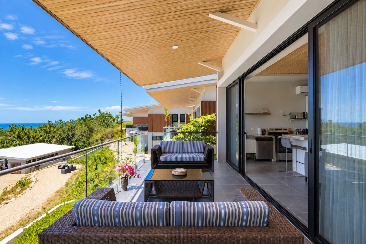 Un espacio exterior moderno con una sala, una mesa de comedor y una cocina visible a través de un ventanal.