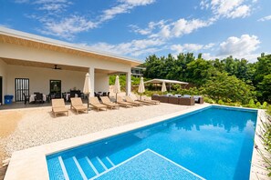 Zona de piscina con sillones de descanso y sombrillas, rodeada de follaje.