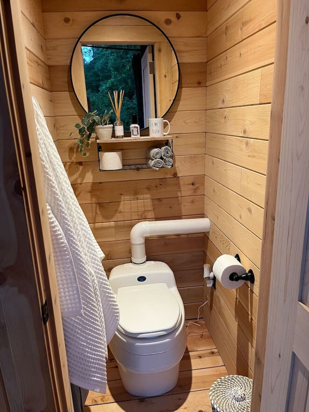Un baño de madera pequeño y funcional con inodoro, un estante con artículos de tocador, una toalla colgada en la pared y una ventana con vistas a árboles.