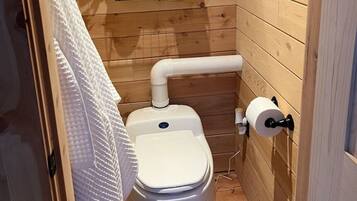 Un baño de madera pequeño y funcional con inodoro, un estante con artículos de tocador, una toalla colgada en la pared y una ventana con vistas a árboles.