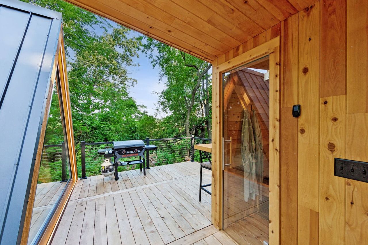 Una terraza de madera con una parrilla y una mesa, una puerta de cristal que da a una habitación y vistas a árboles.