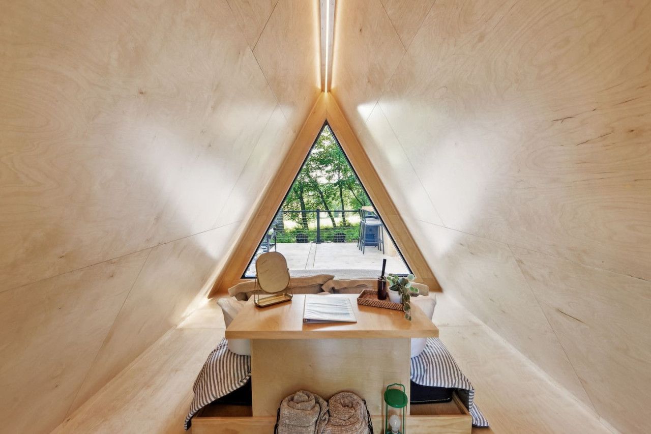 A cozy, triangular wooden tent with a table, a chair, and a view of greenery outside.