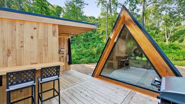 A wooden cabin with a triangular glass extension, a deck with two chairs, and a grill. - New A-Frame Tiny Home Retreat -With Private Sauna! (Christiana)