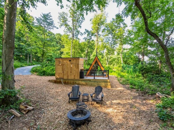 A wooden cabin with a deck, surrounded by a forest. There are two Adirondack chairs and a fire pit in front.
