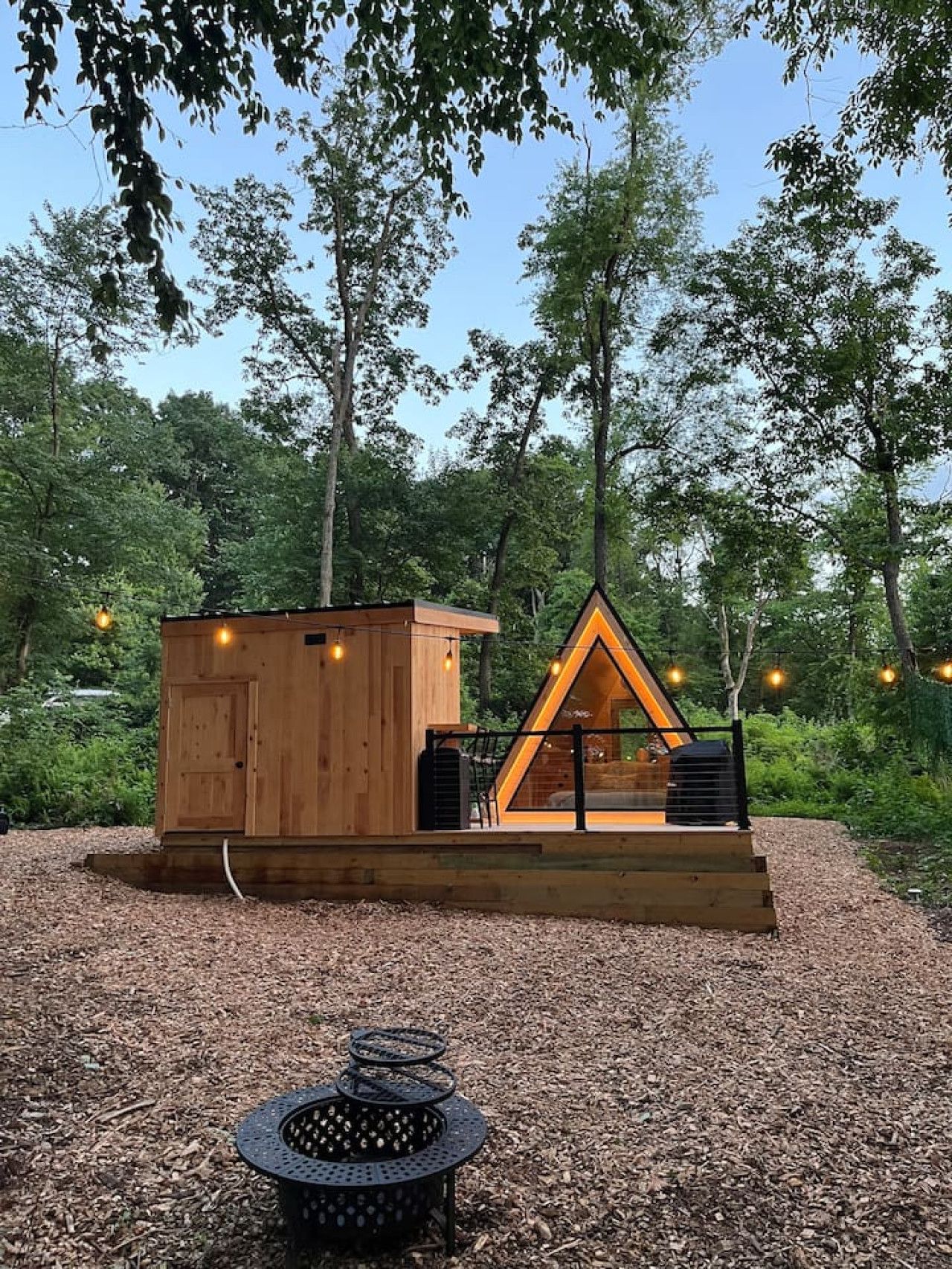 A wooden cabin with a triangular extension, surrounded by trees and a gravel area with a fire pit.