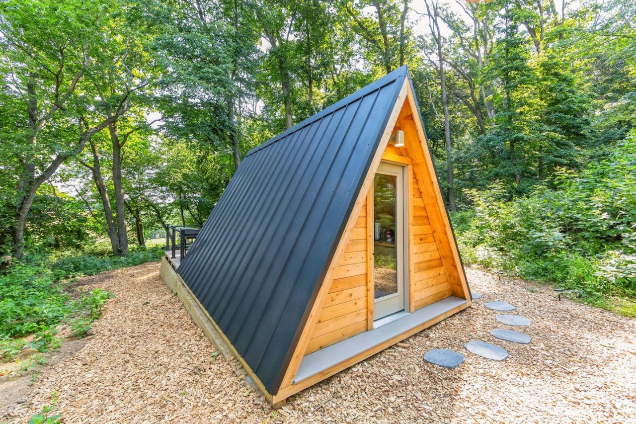 Una pequeña cabaña de madera triangular con techo oscuro, rodeada de un bosque.