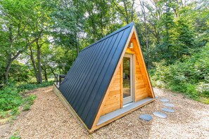 A small, triangular wooden cabin with a dark roof, surrounded by a forest.