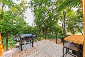 A wooden deck with a grill, a table, and chairs, surrounded by trees. - New A-Frame Tiny Home Retreat -With Private Sauna! (Christiana)