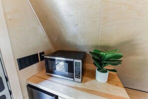A compact kitchen with a microwave on a wooden countertop, a potted plant, and a wall-mounted electrical outlet. - New A-Frame Tiny Home Retreat -With Private Sauna! (Christiana)