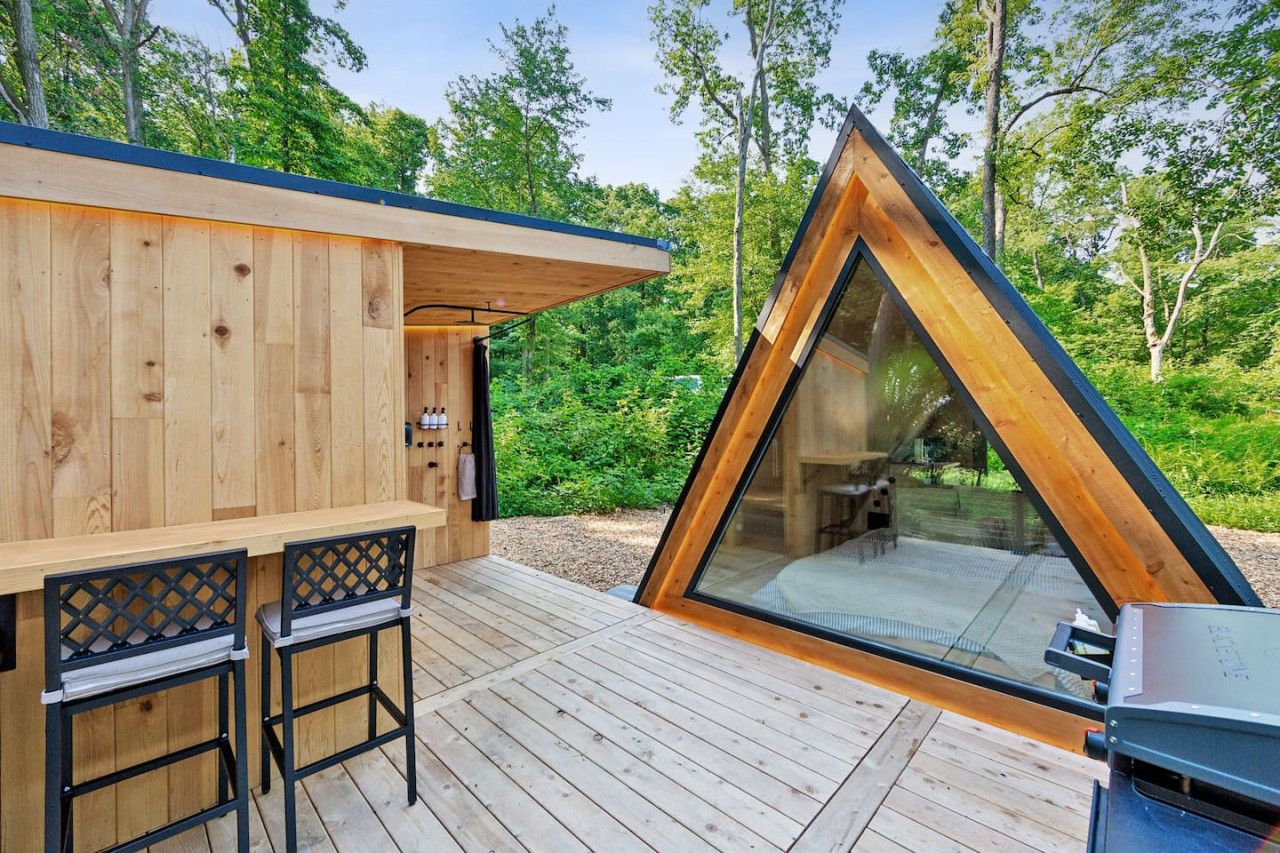 Una cabaña de madera con un porche triangular de vidrio, una terraza con dos sillas y una parrilla.